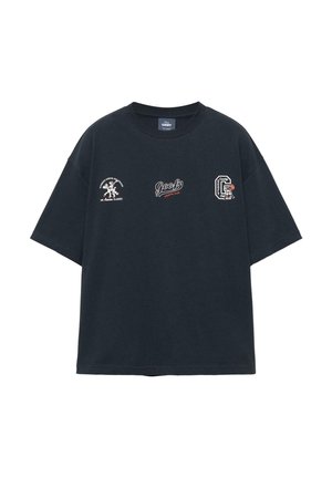 T-shirt noir surdimensionné avec trois petits logos à thème Goofy disposés sur la poitrine, comprenant des textes et des dessins de personnage de dessin animé.
