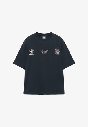 T-shirt noir surdimensionné avec trois petits logos à thème Goofy disposés sur la poitrine, comprenant des textes et des dessins de personnage de dessin animé.