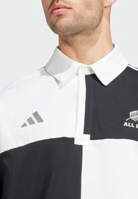 Svart och vit polo skjorta med strukturerad krage, som har knappstängning, texturerat tyg och silverfärgad Adidas-logotyp på vänster sida.