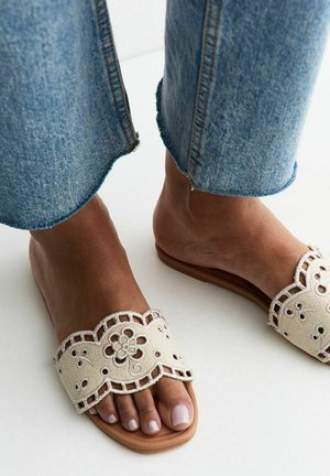 Voeten met blauwe spijkerbroek en beige open sandalen met bloemige uitgestanste borduursels op een witte achtergrond.