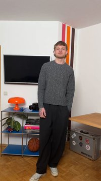 Jeune homme en pull gris et pantalon noir se tient dans le salon avec un ballon de basket, une caméra, une lampe orange et une valise décorée d'autocollants.