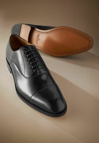 Zwarte leren dresschoenen met een slanke, spitse neus, een gesloten vetersysteem en een tan leren zool met zichtbare stiksels.