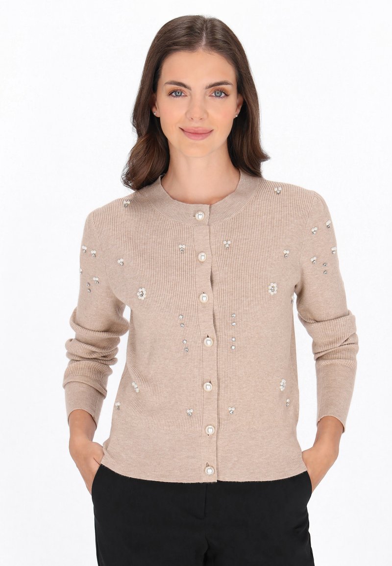 Beige ribbad kavaj med rund halsringning, prydd med dekorativa kulor och blommönster, med knappfront och långa ärmar.