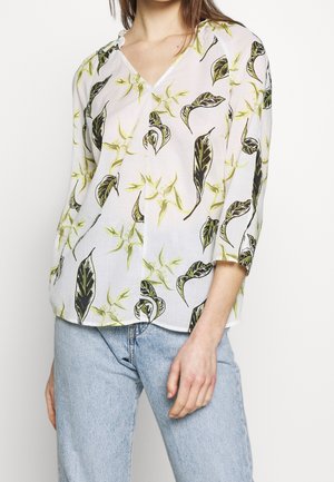 Woman wearing white V-neck blouse with green and black leaf pattern and light blue jeans, standing against a plain background.