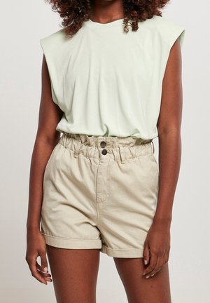 Frau trägt ein hellgrünes ärmelloses Oberteil und beige Paperbag-Shorts mit hohem Bund und umgeschlagenen Säumen vor schlichtem Hintergrund.