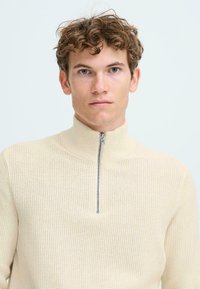Lichtbeige gebreide zip-up sweater met een hoge kraag en zilveren ritsdetails, met een gestructureerd patroon in de stof.