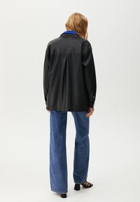 Kvinna står vänd bortåt iförd en svart oversized jacka med en blå krage, vida blå jeans och svarta klacksandaler mot en vit bakgrund.