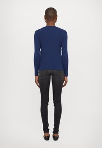 Mörkblå långärmad topp med ribbad textur, lätt rundad nederkant, i kombination med mörka jeans och svarta ballerina-skor, sedd bakifrån.