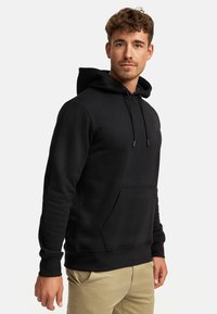 Mężczyzna z kręconymi włosami, ubrany w czarną bluzę z kapturem i sznurkami oraz beżowe spodnie, stojący na tle jednolitego jasnego tła.