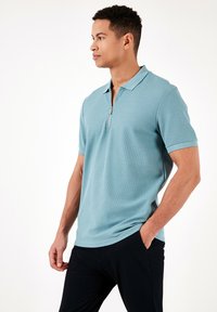 Gaiši zils īsām piedurknēm polo krekls ar rāvējslēdzēja kakla izgriezumu un teksturētu audumu. Iespējo klasisku apkakli un brīvu piegriezumu.