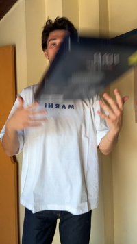 Objeto retangular preto sendo lançado, com uma t-shirt branca com a inscrição "MARIN" em letras azuis. O fundo mostra uma parede de cor clara.