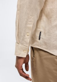 Μανσέτα μπεζ μακρυμάνικου λινού πουκαμίσου με δύο κουμπιά, φορεμένη από ένα άτομο, εμφανίζοντας μέρος από τα ταιριαστά μπεζ παντελόνια και μια μικρή ετικέτα ενδύματος.