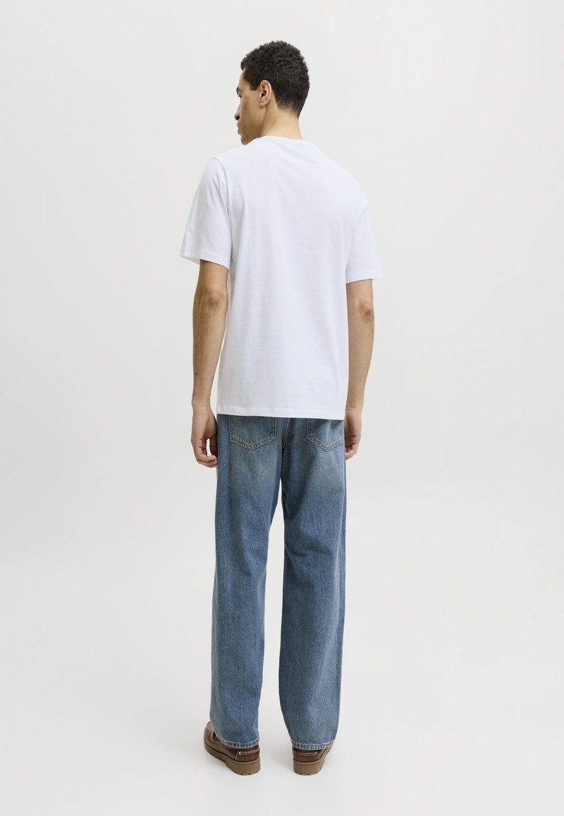 Hombre que lleva una camiseta blanca lisa, vaqueros azules y zapatos marrones, de pie con la espalda hacia la cámara sobre un fondo blanco liso.
