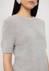 Pull en maille gris clair à manches courtes, avec un col rond et une finition texturée, présentant des détails de couture visibles.