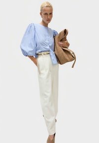 Jasnoniebieska bluzka w paski z bufkami, białe spodnie z wysokim stanem, torebka z plecionki w kolorze koniakowym oraz beżowe szpilki. Neutralne tło.