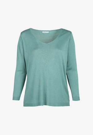 Kvinde med krøllet hår iført grønblå bluse med lange ærmer og grå jeans, hænderne i lommerne, beskåret fra skuldre til hofter.
