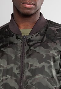 Uomo che indossa una giacca a zip camouflage verde scuro sopra una camicia verde oliva, bocca e parte bassa del viso visibili, sfondo neutro.