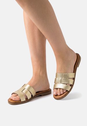 Voeten met metalen gouden instapsandalen met brede banden en platte bruine zolen op een eenvoudige lichte achtergrond.