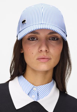 Nő kék-fehér csíkos sapkával kitűzővel, hozzáillő csíkos inggallérral és fekete ruhadarabbal fehér gallérral.