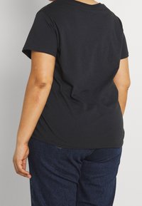 Persoon met de rug naar ons toe, gekleed in een eenvoudige zwarte T-shirt met korte mouwen en een donkere spijkerbroek tegen een eentonige lichte achtergrond.