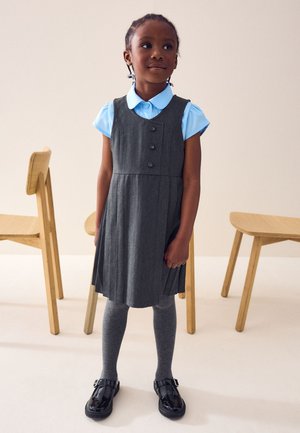 Ragazza giovane in piedi al chiuso, indossa un grembiule scolastico grigio plissettato, una camicia a maniche corte azzurro chiaro, collant grigi e scarpe nere con fibbia.