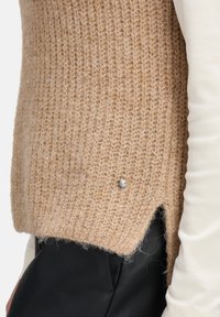 Beżowy sweter z dzianiny o strukturalnym wykończeniu i srebrnym guzikem z boku. Posiada rozcięty dół i jest zestawiony z białą koszulą.