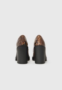 Καφέ παπούτσια με σχέδιο φιδιού και ψηλό τακούνι, με υφή στην επιφάνεια, στρογγυλή μύτη και στιβαρό μαύρο τετράγωνο τακούνι. Όψη από πίσω σε λευκό φόντο.