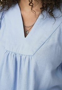 Blusa in lino azzurro chiaro con scollatura a V e dettagli arricciati sotto il busto. Presenta una texture morbida e accenti di cucitura sottili.