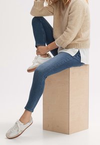 Béžový vlněný svetr, pod ním bílá košile, modré džíny a bílé tenisky s hnědými podrážkami, sedící na kostce z jasného dřeva.