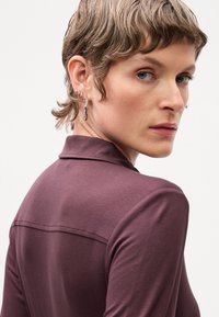 Haut à manches longues violet avec un col, présentant une texture lisse et une couture horizontale dans le dos. L'oreille du modèle est ornée de plusieurs boucles d'oreilles en argent.