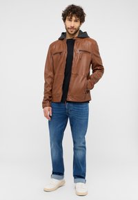 Braune Lederjacke mit Reißverschlusstaschen, kombiniert mit einem grauen Kapuzenfutter. Getragen über einem schwarzen Hemd, kombiniert mit blauen Jeans und weißen Sneakern.