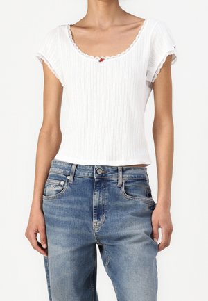 Hvid ribbet kortærmet top med bølgede kanter og lille rød knapdetalje, kombineret med falmede blå denimjeans.