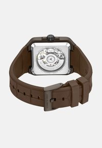 Vierkante roestvrijstalen horloge met een transparante achterkant, bruine siliconen band en zichtbaar zilveren mechanisch uurwerk aan de binnenkant.