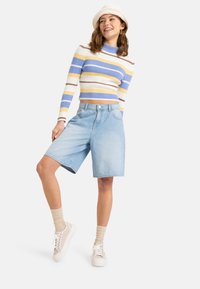 Helle blaue Jeans-Shorts mit lockerer Passform, kombiniert mit einem kurz geschnittenen, gestreiften Pullover in Blau, Gelb und Braun. Weiße Sneaker und beigefarbene Socken.