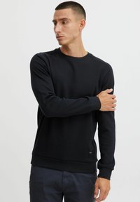Sweatshirt noir à col rond en tissu texturé, avec des poignets et un ourlet côtelés. Présente une petite étiquette de logo sur le côté inférieur.