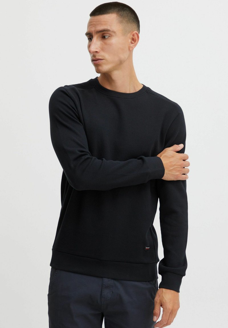Sweatshirt noir à col rond en tissu texturé, avec des poignets et un ourlet côtelés. Présente une petite étiquette de logo sur le côté inférieur.