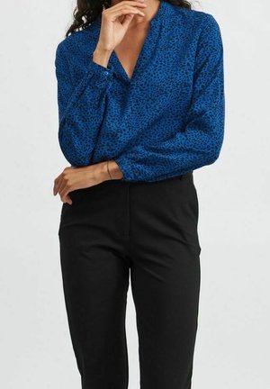 Kvinde iført en blå langærmet bluse med sorte pletter, stoppet ned i højtaljede sorte bukser, hånd hvilende nær hendes hage.