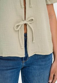 Lyse beige knappeskyder lavet af tekstureret stof, med en frontbinding, korte ærmer og en let cropped kant. Kombineret med blå jeans.