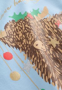 Jasnoniebieska tkanina z brązowym, fakturowanym motywem niedźwiedzia z zielonym ostrokrzewem, czerwonymi i złotymi ozdobami oraz metalicznymi akcentami w zabawnym układzie.