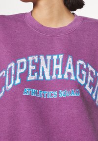 Fialová mikina se žebrovaným výstřihem, na které je velkým, opotřebovaným bílým a modrým písmem nápis "COPENHAGEN" a pod ním "ATHLETICS SQUAD".
