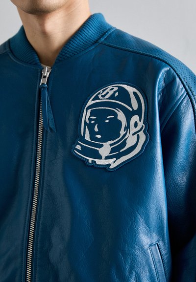 Niebieska skórzana kurtka bombowiec z srebrną naszywką astronauty, prążkowanym kołnierzem i szczegółowym zamkiem błyskawicznym. Gładka tekstura z boczną kieszenią.