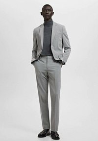 Szary garnitur na miarę z dopasowaną marynarką i spodniami z płaskim przodem, zestawiony z ciemnym golfem. Gładka tkanina i klasyczny krój.