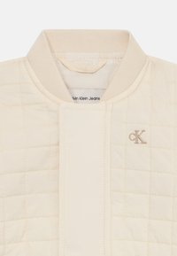 Veste matelassée de couleur crème avec un col côtelé et le logo CK. Le tissu semble lisse avec des motifs de couture horizontaux et verticaux.