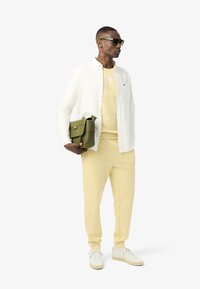 Sudadera y joggers amarillos claros, camisa blanca de botones, bolso verde oliva y zapatillas blancas, con gafas de sol negras como acento.