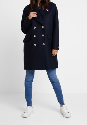 Kvinde iført en dobbeltradet navyfrakke med sølvknapper, blå skinny jeans og hvide sneakers, stående med benene spredt.