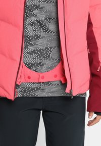 Personne portant une veste rose par-dessus une chemise à motifs gris et noir, avec un pantalon noir, et une main partiellement visible sur le côté.