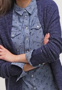 Marinblå stickad cardigan över en ljusblå, knappad chambrayskjorta med strukturerat mönster och två bröstfickor.