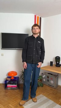 Melna krekla ar kontrastējošiem baltajiem šuvumiem, divas krūšu kabatas, zila džinsu bikses un brūnas klogs, iestatīts mūsdienīgā interjerā.