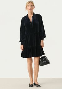 Rochie neagră din catifea cu decolteu în V, mâneci lungi tip puff și fustă stratificată. Purtați cu o poșetă neagră și pantofi cu buline. Textură netedă.