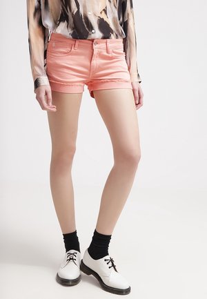 Persoon draagt perzikkleurige denimshorts met rafelige zomen, zwarte sokken, witte veter schoenen en een lange mouwen patroon overhemd met knopen.